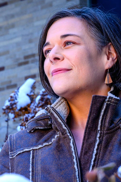 A woman with short dark hair smiles slightly while looking off-camera. She wears a brown, fleece-lined winter jacket. In the background, snow rests on dried plants in front of a tan brick wall.